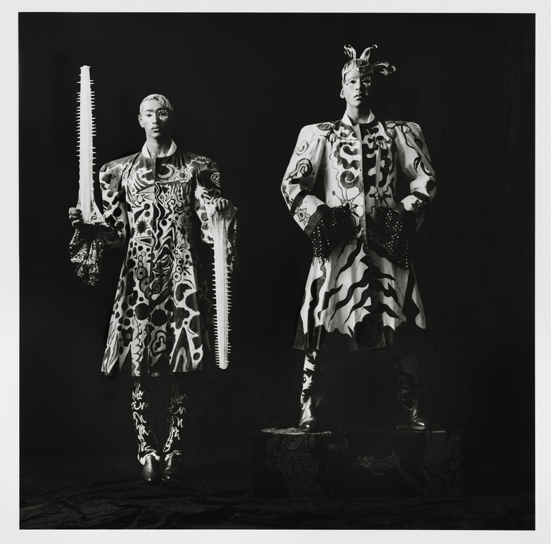Two figures in patterned coats pose in a studio portrait; one holds a saw. The photograph is black and white with sharp focus.