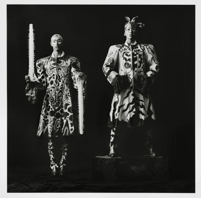 Two figures in patterned coats pose in a studio portrait; one holds a saw. The photograph is black and white with sharp focus.