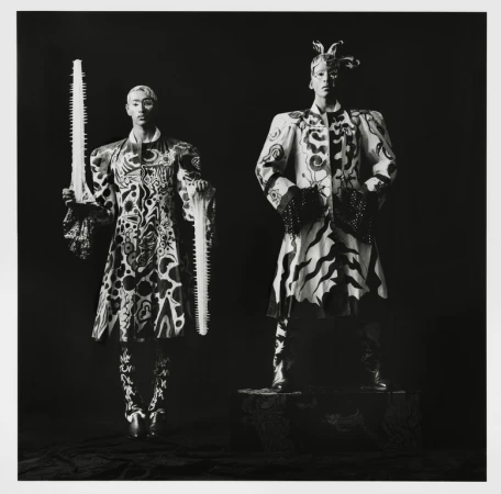 Two figures in patterned coats pose in a studio portrait; one holds a saw. The photograph is black and white with sharp focus.