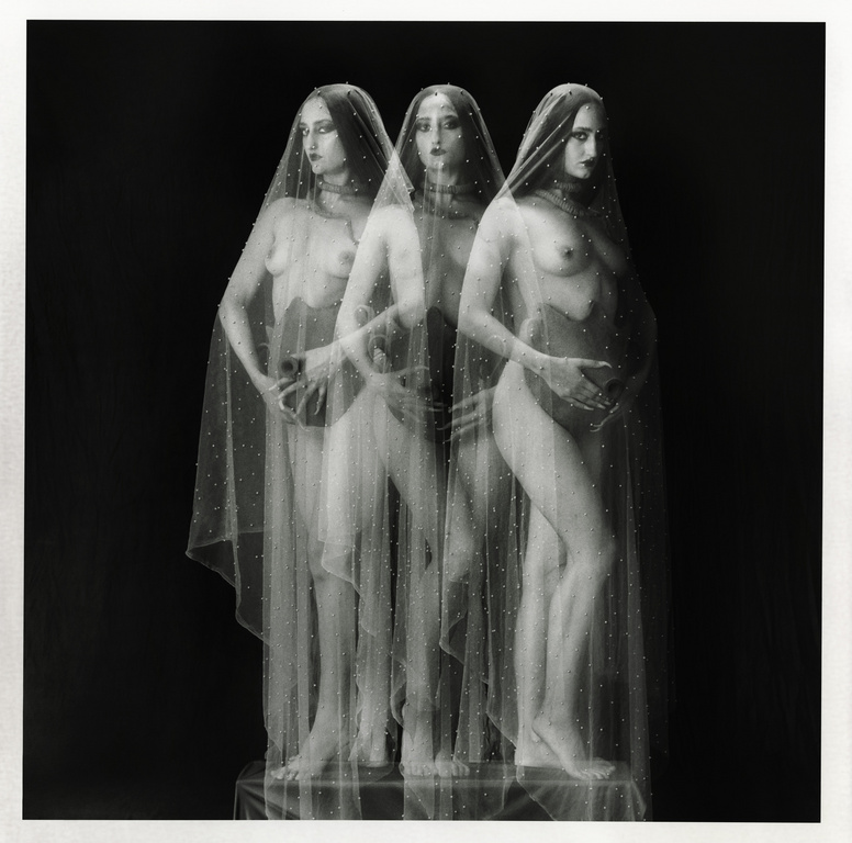 Three pregnant women stand in a dark studio. The women are partially obscured by a thin, spotted translucent veil. The photo is black and white.