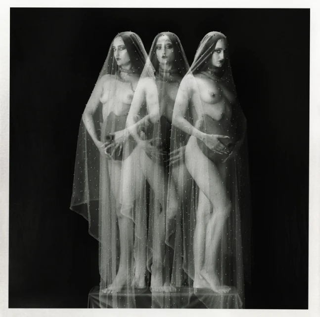 Three pregnant women stand in a dark studio. The women are partially obscured by a thin, spotted translucent veil. The photo is black and white.