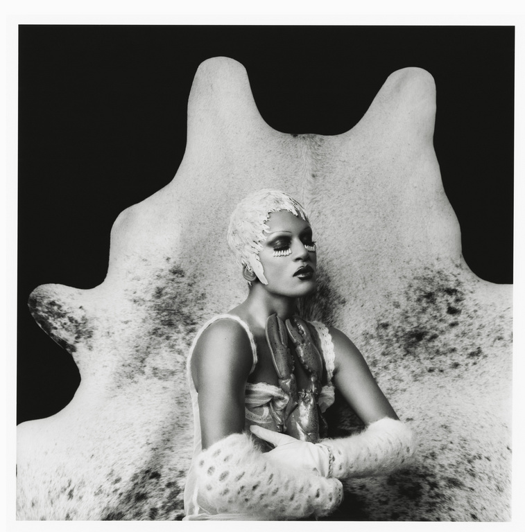 Monochrome portrait of a person with elaborate face makeup, decorative head covering, and fur accessories, posed in front of a hide.