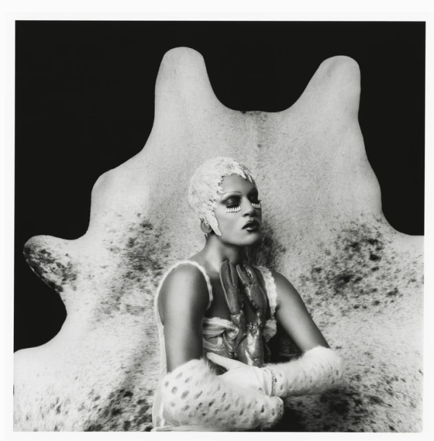 Monochrome portrait of a person with elaborate face makeup, decorative head covering, and fur accessories, posed in front of a hide.