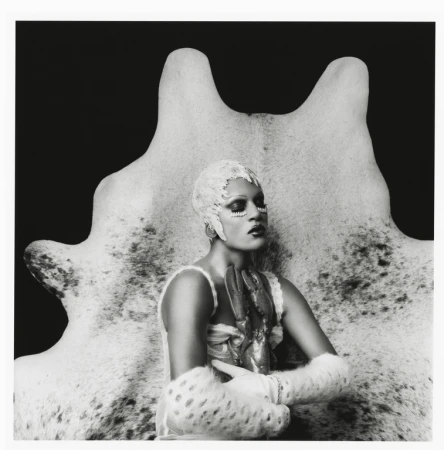Monochrome portrait of a person with elaborate face makeup, decorative head covering, and fur accessories, posed in front of a hide.