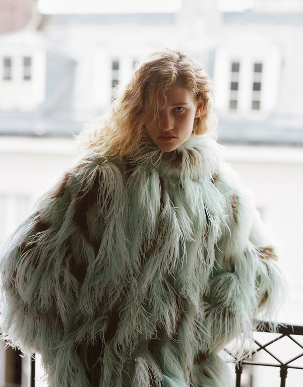 Fashion shot of a blonde woman in a feathery light blue coat standing on a balcony, with city buildings in the background.