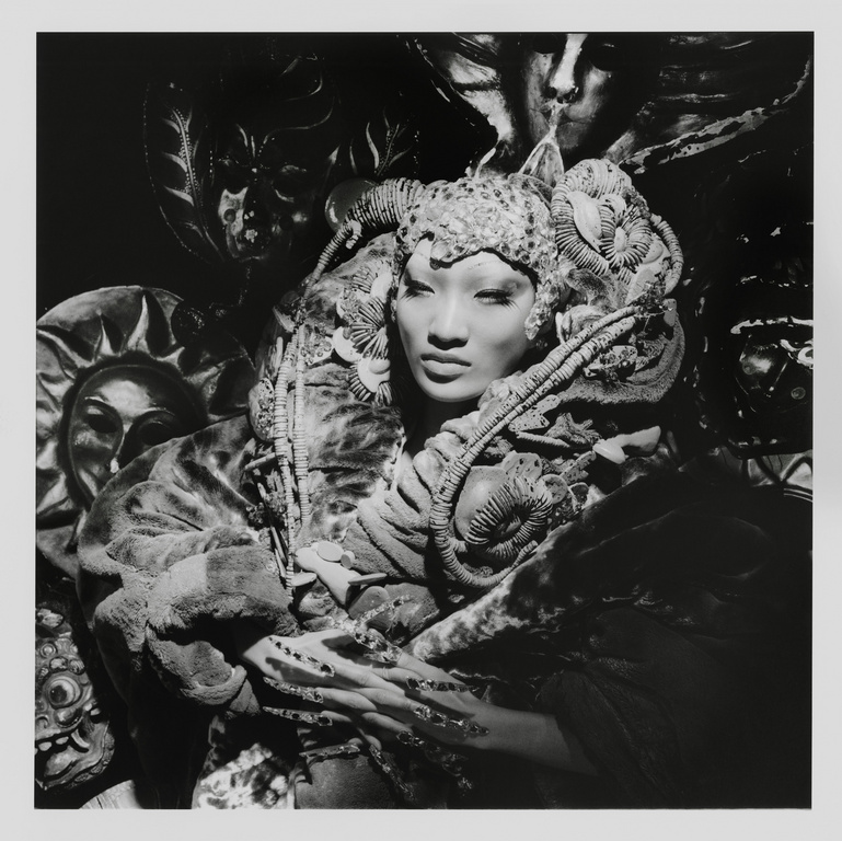 Striking monochrome portrait of an Asian woman with elaborate headwear and makeup surrounded by artistic masks, creating a surreal visual.