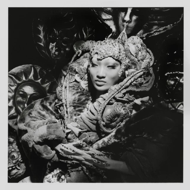 Striking monochrome portrait of an Asian woman with elaborate headwear and makeup surrounded by artistic masks, creating a surreal visual.
