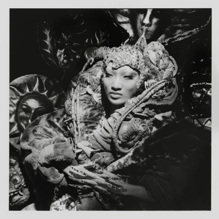 Striking monochrome portrait of an Asian woman with elaborate headwear and makeup surrounded by artistic masks, creating a surreal visual.