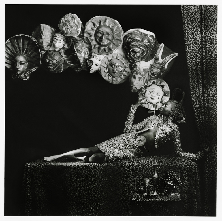 Monochrome image of a person in a sequined dress reclining on a patterned surface, holding a mask, surrounded by other masks.