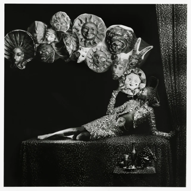 Monochrome image of a person in a sequined dress reclining on a patterned surface, holding a mask, surrounded by other masks.