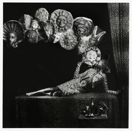 Monochrome image of a person in a sequined dress reclining on a patterned surface, holding a mask, surrounded by other masks.