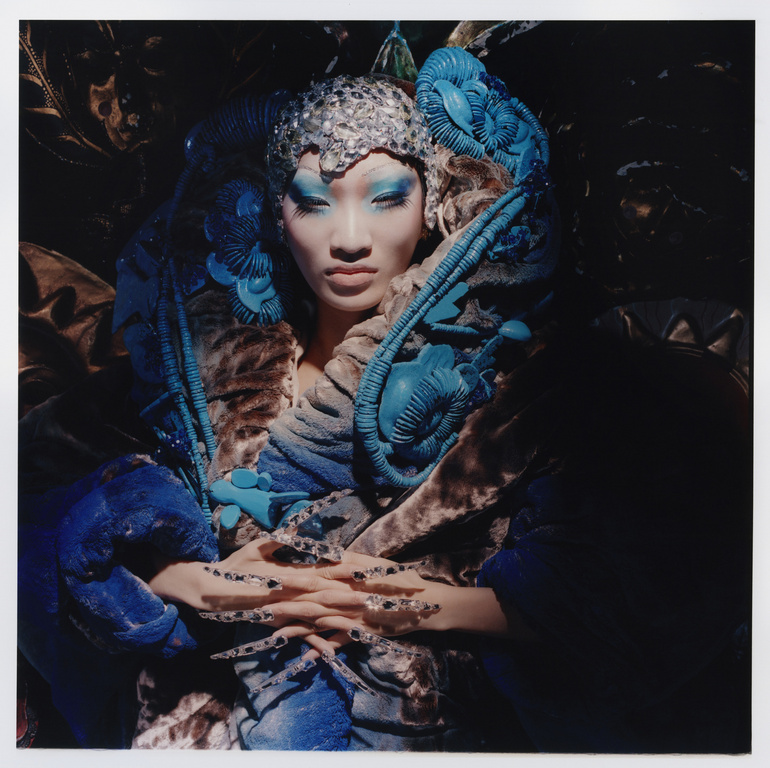 A person with dramatic makeup and an elaborate turquoise and silver headpiece is photographed in a dimly lit studio with neutral background.