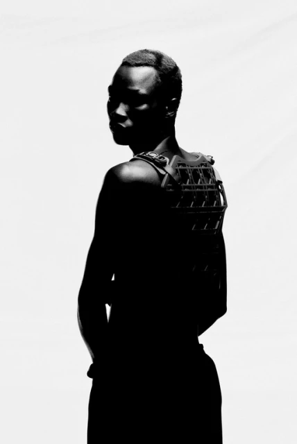 Silhouette of a Black male with short hair, wearing an elaborate back brace or harness, against a stark white background in studio lighting.