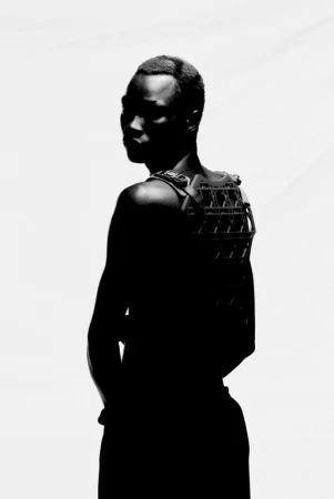 Silhouette of a Black male with short hair, wearing an elaborate back brace or harness, against a stark white background in studio lighting.