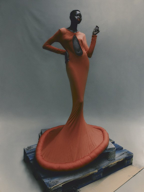 A Black woman stands on a wooden pallet in a bodycon dress with a large central cutout, set against a light gray backdrop.