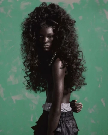 Black woman with voluminous curly hair wearing dark shorts against a textured green background, cast by Affa Osman.