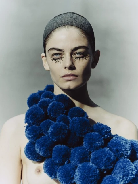 Close-up portrait of a young woman with creative eye makeup and blue pompoms draped over her shoulder, against a neutral grey background.