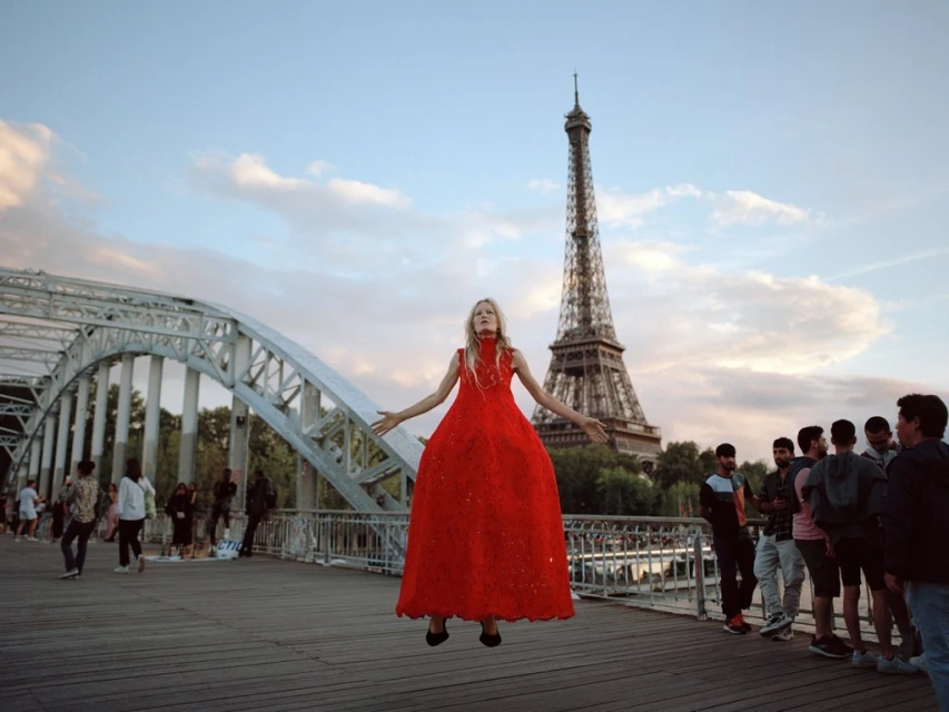 Woman in red dress poses on bridge with Eiffel Tower in the background. She has blonde hair and is surrounded by a crowd.