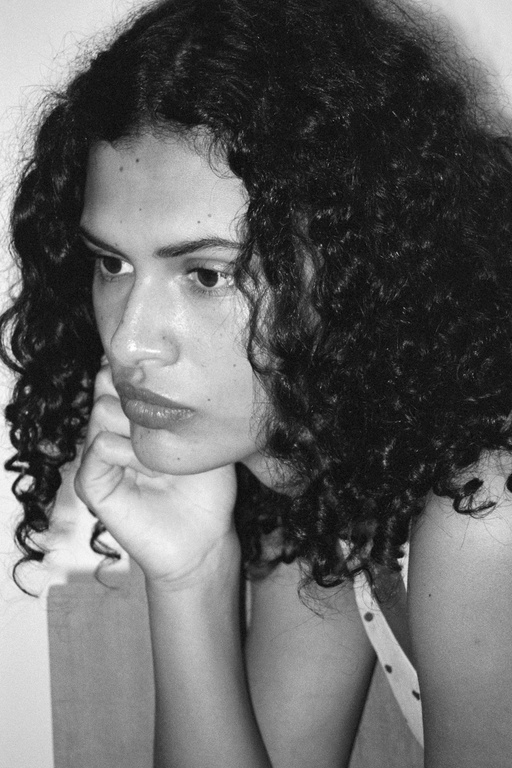 Portrait of a pensive adult woman with curly hair, resting her chin on her hand, wearing a polka dot top in a bright, indoor setting.