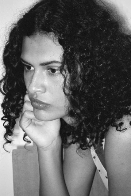 Portrait of a pensive adult woman with curly hair, resting her chin on her hand, wearing a polka dot top in a bright, indoor setting.