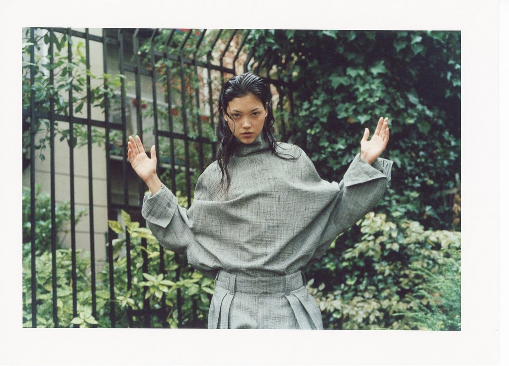 Asian woman in grey oversized shirt and pleated skirt poses in front of a wrought iron fence with greenery in an outdoor fashion shoot.