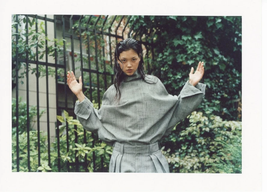 Asian woman in grey oversized shirt and pleated skirt poses in front of a wrought iron fence with greenery in an outdoor fashion shoot.