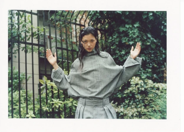 Asian woman in grey oversized shirt and pleated skirt poses in front of a wrought iron fence with greenery in an outdoor fashion shoot.
