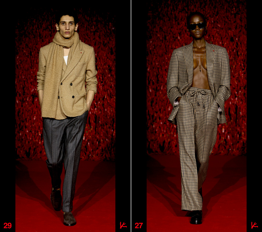 Two fashion models walk the runway during a show, one in a tan suit and the other in a brown checkered suit against a red backdrop.