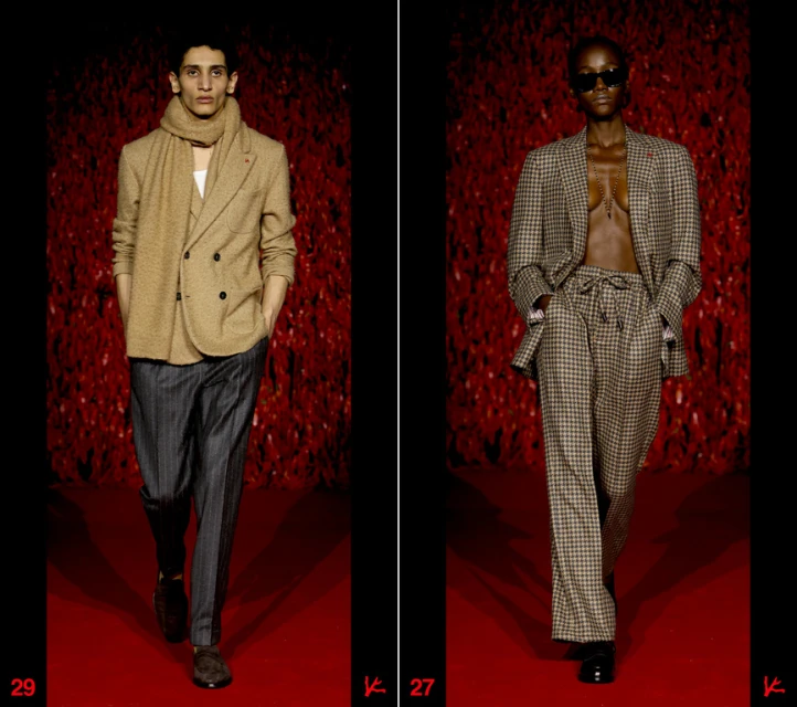 Two fashion models walk the runway during a show, one in a tan suit and the other in a brown checkered suit against a red backdrop.
