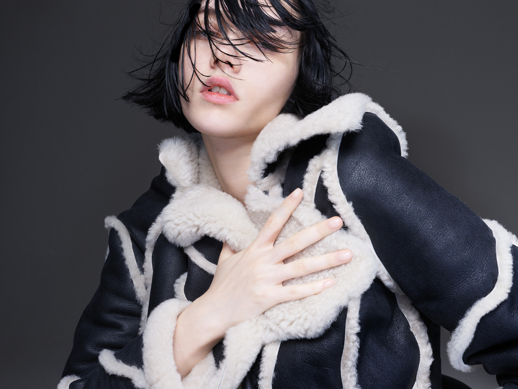 Close-up portrait of a woman with dark, wet hair wearing a black sheepskin coat against a muted gray backdrop, fashion style.