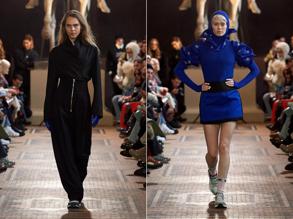 Two fashion models in runway show. Model on the left wears black top with pants, model on right in blue mini dress with hood.