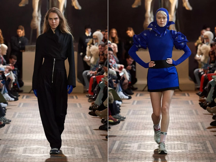 Two fashion models in runway show. Model on the left wears black top with pants, model on right in blue mini dress with hood.