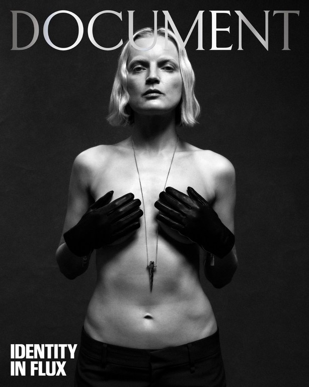 Black and white portrait of a shirtless fair-skinned woman with blonde hair wearing black gloves and a pendant against a dark background.