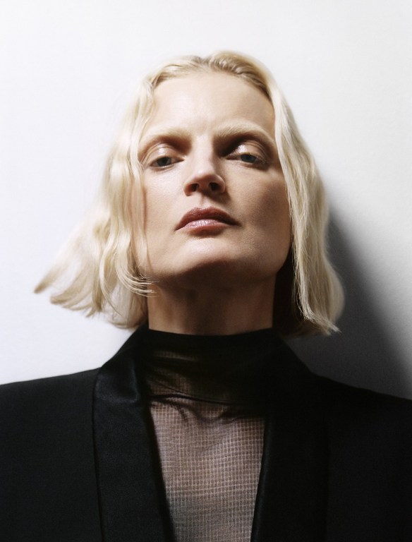 Close-up portrait of a blonde woman in a black blazer and sheer top against a white backdrop, showcasing fashion and beauty.