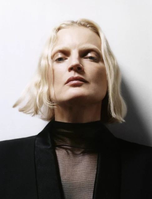 Close-up portrait of a blonde woman in a black blazer and sheer top against a white backdrop, showcasing fashion and beauty.
