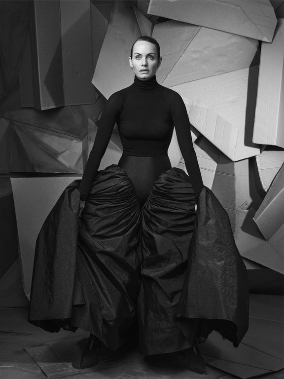 Elegant adult female posing in a dark dress with exaggerated hip puffs against a geometric cardboard background in black and white.