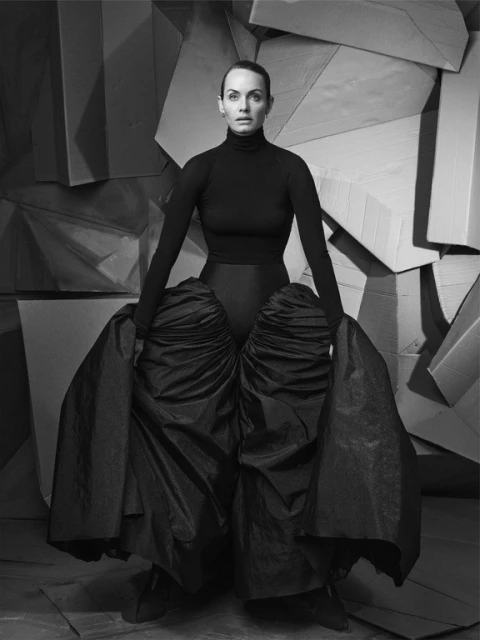 Elegant adult female posing in a dark dress with exaggerated hip puffs against a geometric cardboard background in black and white.