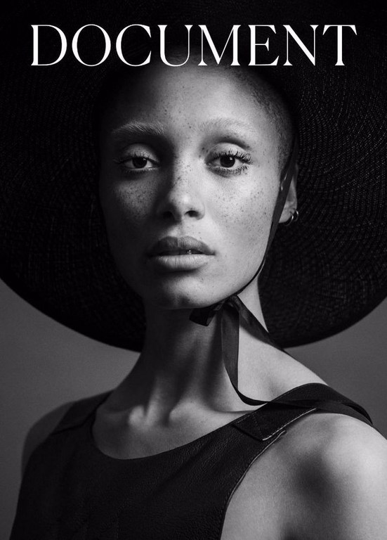 Black and white portrait of a woman with shaved head wearing a large hat and a dark top against a neutral background.