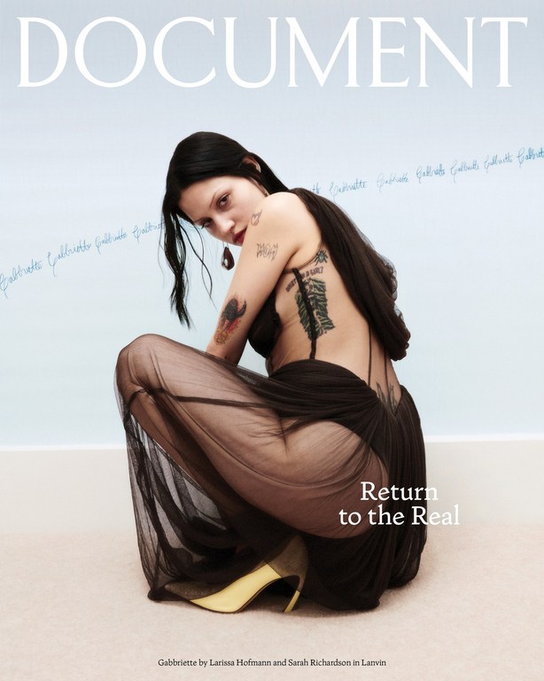 Magazine cover featuring a crouched young woman with tattoos wearing a sheer black dress and yellow heels against a light backdrop.