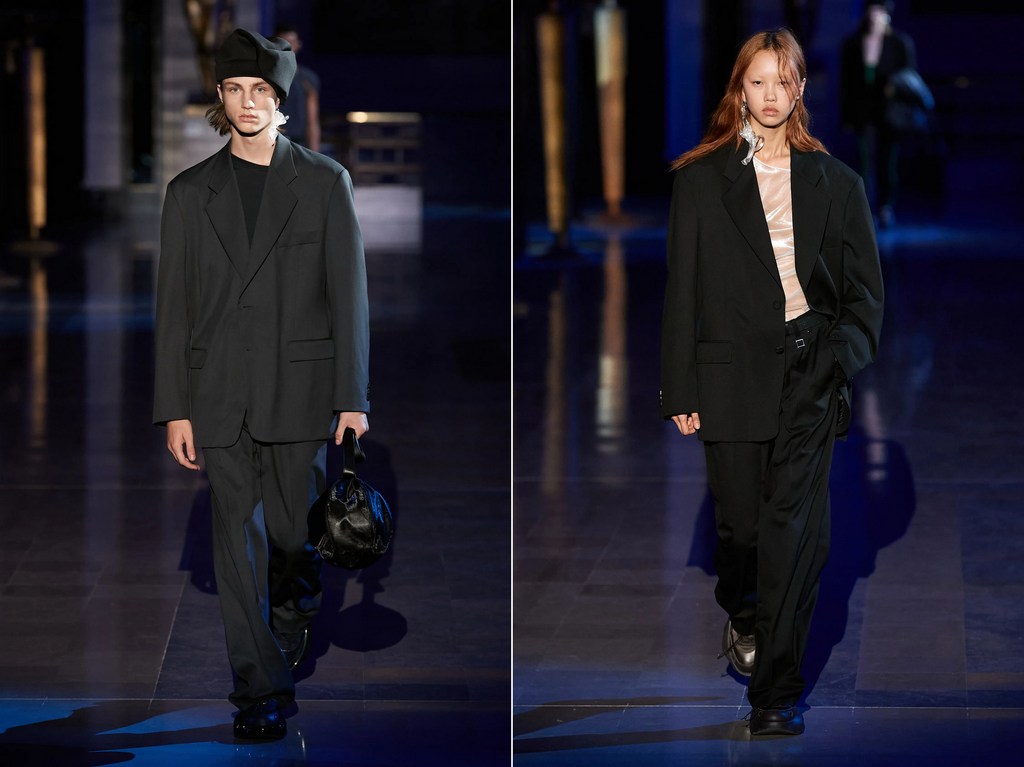 Two fashion models present dark, oversized menswear on a runway, featuring long suit jackets, wide trousers, and bucket hats, in a dimly lit setting.