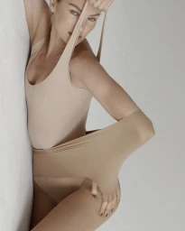 Model wearing a nude bodysuit and tights against a seamless white background, holding and stretching the fabric.