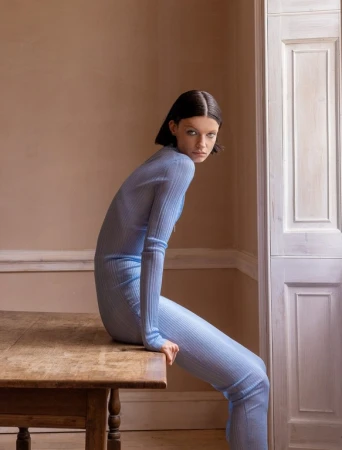 Model in powder blue ribbed bodysuit seated on a wooden table in a room