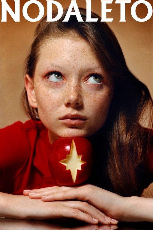 Portrait of a young woman with freckles looking upwards, posed with her chin resting on a red apple with a star carved into it.