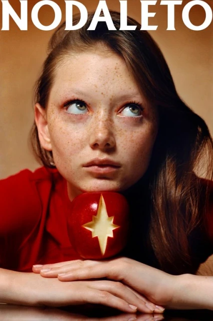 Portrait of a young woman with freckles looking upwards, posed with her chin resting on a red apple with a star carved into it.