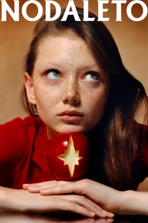 Portrait of a young woman with freckles looking upwards, posed with her chin resting on a red apple with a star carved into it.