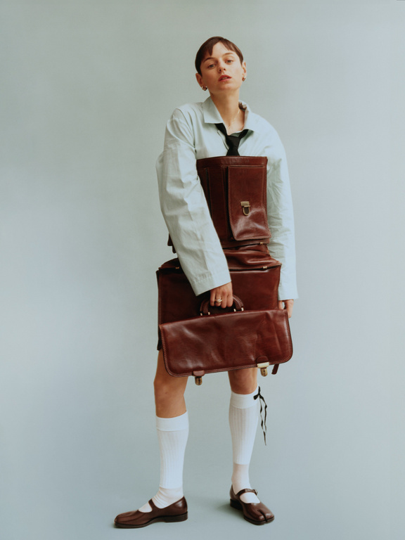A woman posing with a stack of brown leather briefcases. She is wearing a blue shirt and white socks against a light blue background.