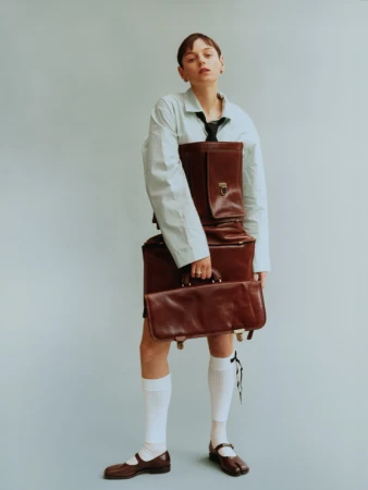 A woman posing with a stack of brown leather briefcases. She is wearing a blue shirt and white socks against a light blue background.