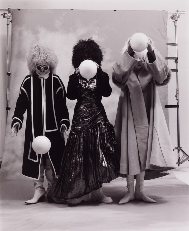 Three people in black-and-white studio shot. One in metallic gown and two in coats with fur, holding white balls, fashion shoot.