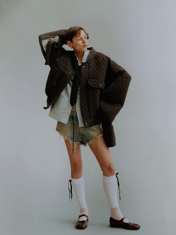 A woman in a dark brown quilted jacket, light blue shirt, and denim shorts poses indoors against a grey background in a fashion editorial.