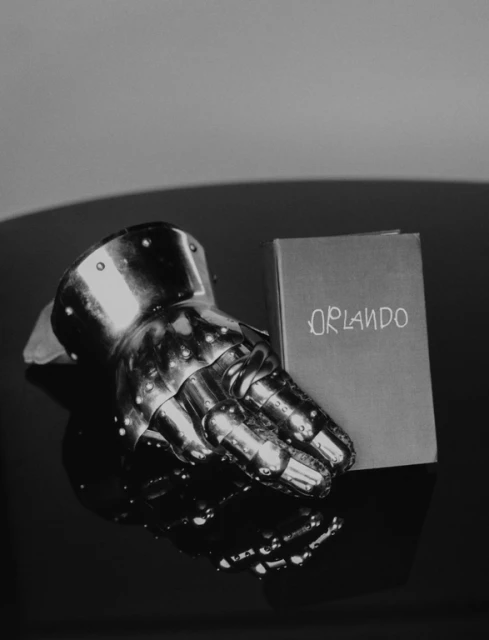 Close-up studio shot of a metal gauntlet resting next to a book titled 'Orlando' on a reflective black surface, creating a dramatic still life.